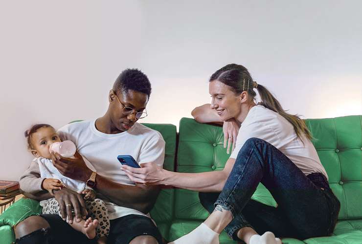 Couple on sofa feed a baby while checking their phone bill