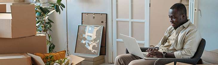 African American man on laptop surrounded by moving boxes sets up his new internet service